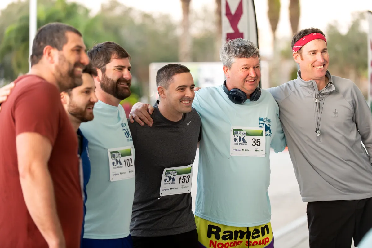 A group of smiling Health in Action 5K participants in race bibs posing with arms around each other.