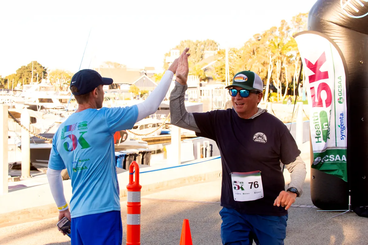Two participants high-five at the Health in Action 5K finish line beside a Syngenta and GCSAA banner.