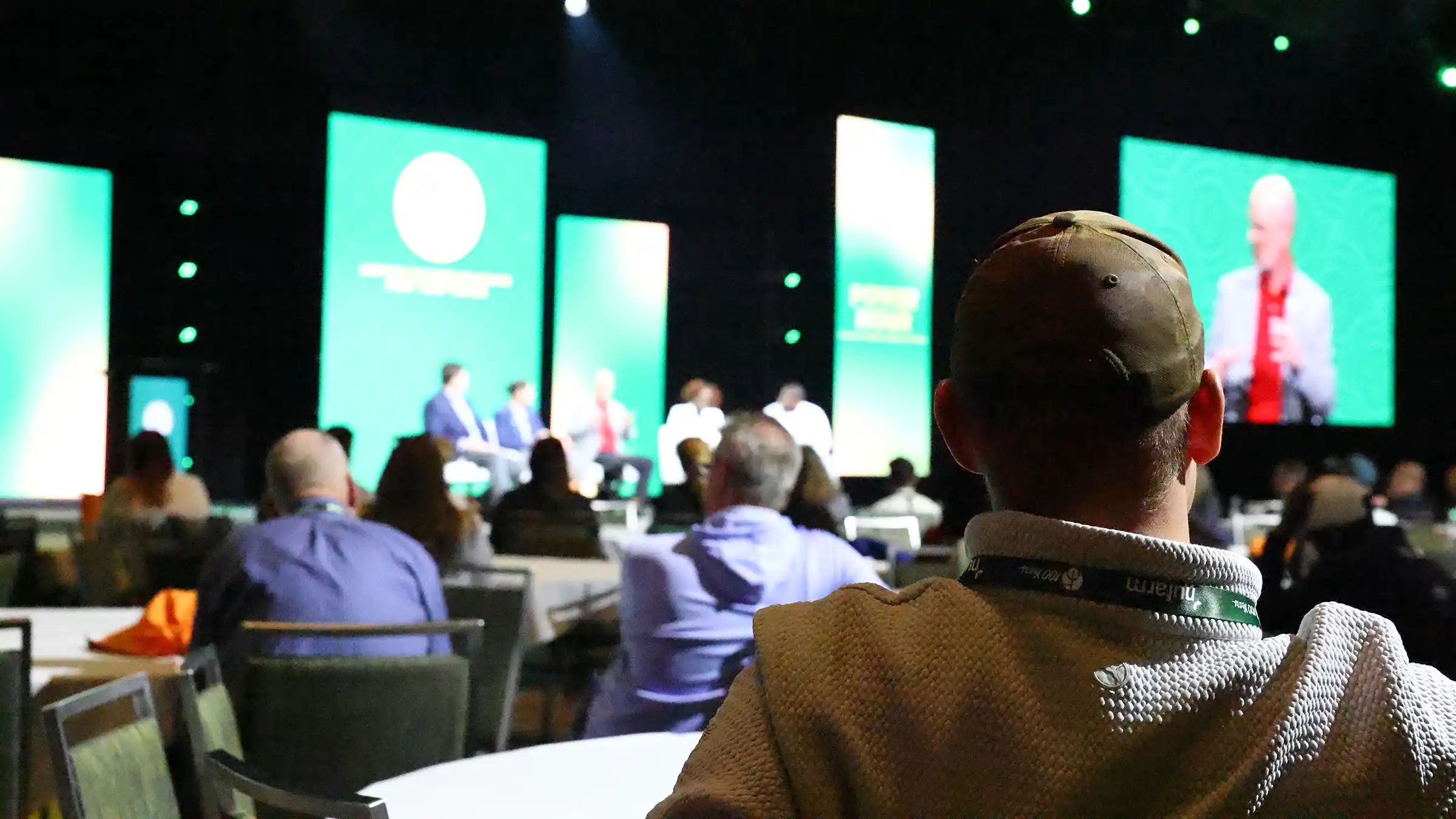 An audience seated at tables watching a panel discussion on a stage with large screens.